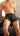 A shirtless man wearing black boxer briefs with a small multicolor geometric pattern, sitting on a black leather couch.